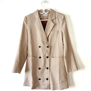 May include: A double-breasted blazer in a tan and white houndstooth pattern featuring notched lapels, long sleeves, and eight dark buttons arranged in two vertical rows. The interior label displays the brand name LIONESS in black text.
