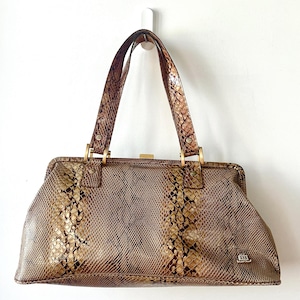 May include: A structured handbag featuring a brown and tan snakeskin-patterned texture. The bag has two top handles attached with gold-tone metal hardware, a rectangular frame closure, and a small metallic logo plaque on the lower right corner.