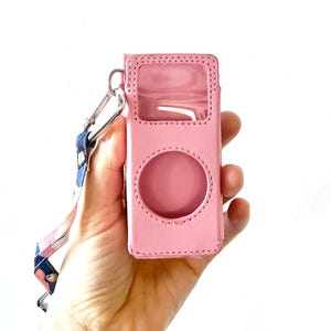 May include: A light pink rectangular protective case featuring a clear plastic window at the top and a circular cutout in the centre. The case includes a silver metal carabiner attached to a blue and pink patterned fabric strap with a metal snap.