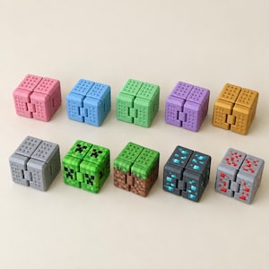 May include: A collection of ten square fidget cubes arranged in two rows. The cubes feature various colors and pixelated textures, including solid pink, blue, green, purple, gold, gray, and patterned designs resembling game blocks.
