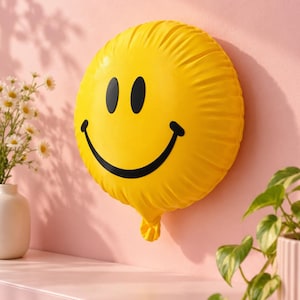 May include: A round, bright yellow balloon featuring a black smiley face graphic with two oval eyes and a wide, curved mouth. The balloon has a gathered, knotted tail at the bottom and a smooth, slightly reflective surface texture.