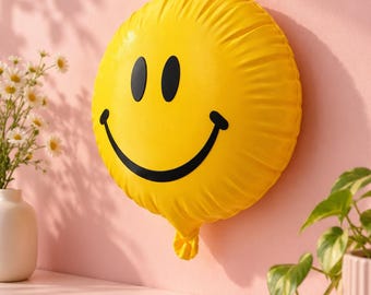 May include: A round, bright yellow balloon featuring a black smiley face graphic with two oval eyes and a wide, curved mouth. The balloon has a gathered, knotted tail at the bottom and a smooth, slightly reflective surface texture.