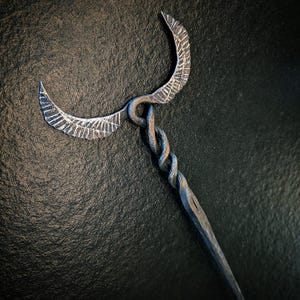 May include: A hand-forged metal hair pin featuring a twisted shaft and a top ornament shaped like two symmetrical, textured wings extending outwards. The metal has a dark, weathered finish with silver highlights along the edges and wing details.