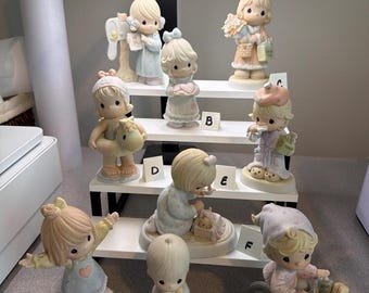 Może przedstawiać: Kolekcja dziewięciu porcelanowych figurek przedstawiających dzieci z oczami w kształcie łez, w pastelowych ubraniach i z różnymi akcesoriami. Figurki są ustawione na białym, wielopoziomowym stojaku, a każda z nich jest oznaczona literą od A do I.
