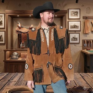 Op de afbeelding: Een beige suède jas met lange zwarte franjes op de borst en zakken, witte stiksels en ronde decoratieve patches op de mouwen. De jas wordt gedragen met een zwarte cowboyhoed en een blauwe spijkerbroek.