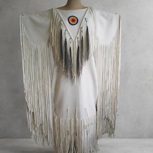 May include: A cream-coloured dress featuring long, flowing fringe along the sleeves and hem. The garment includes a V-shaped neckline adorned with dark grey fringe accents, silver-toned beads, and a circular patch with a blue, yellow, and orange sunburst design.