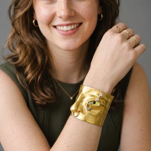 Può includere: Un ampio bracciale a fascia in oro con un design a volto umano scolpito. Il bracciale presenta un naso prominente con un piccolo anello, un singolo occhio con iride verde e pupilla nera, e piccole pietre chiare incastonate sulla superficie.