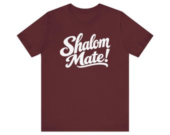 May include: A maroon short-sleeved t-shirt featuring the white text Shalom Mate! printed in a bold, cursive script across the center of the chest. The garment has a crew neckline and a standard fit, showcasing a clean and simple design.