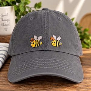 May include: A charcoal gray baseball cap featuring two embroidered bumblebees on the front panel. Each bee is depicted with yellow and black stripes, orange antennae, and light gray wings. The cap has a curved brim and stitched eyelets.
