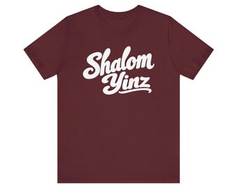 May include: A maroon short-sleeved t-shirt featuring the white text Shalom Yinz printed in a cursive script font across the center of the chest. The garment has a crew neckline and a standard fit, displaying the text clearly against the dark background.