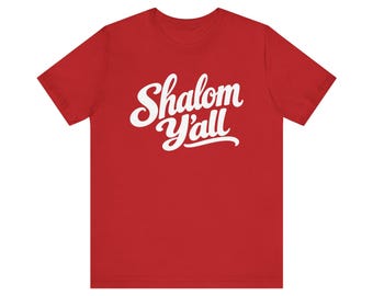 May include: A red short-sleeved t-shirt featuring the white text Shalom Y'all printed in a cursive script font across the center of the chest. The garment has a crew neckline and a standard fit with short sleeves.