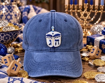 May include: A blue denim baseball cap featuring an embroidered white and blue dreidel design on the front panel. The dreidel embroidery includes Hebrew letters in blue thread, centered on the cap above the curved brim with stitched detailing.