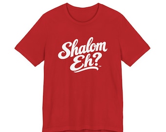 May include: A red short-sleeved t-shirt featuring the white text Shalom Eh? printed in a cursive script font across the center of the chest. The design includes a small trademark symbol located to the right of the word Eh.