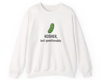 May include: A white long-sleeve crewneck sweatshirt featuring a graphic of a green pickle with a smiling face. Below the pickle, the black text reads: KOSHER, but questionable. The garment has ribbed cuffs and a ribbed hemline.