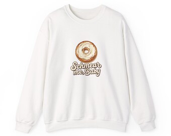 May include: A white long-sleeve crewneck sweatshirt featuring a graphic of a bagel with cream cheese in the center. Below the bagel, the text reads "Schmear me, Baby" in a brown, rounded, retro-style font. The garment has ribbed cuffs.