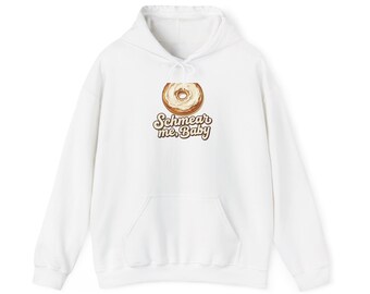 May include: A white hooded sweatshirt featuring a graphic of a bagel with cream cheese and the brown text "Schmear me, Baby" printed underneath. The garment includes a front kangaroo pocket, long sleeves, and a drawstring hood.