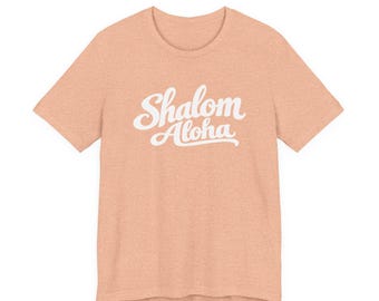 May include: A heathered peach-colored short-sleeve t-shirt featuring the white text Shalom Aloha printed in a cursive script font across the center of the chest. The garment has a crew neckline and a standard fit.