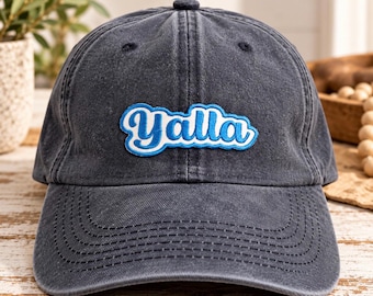 May include: A charcoal gray baseball cap with a distressed, washed-out finish. Centered on the front is an embroidered patch featuring the word Yalla in a white script font with a thick, bright blue outline and a secondary white border.