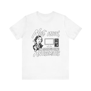 May include: A white short-sleeved t-shirt featuring a black graphic of a person holding a teacup next to a computer. The text reads: Not now, I'M CYBERBULLYING ANTISEMITES. The design is centered on the chest of the garment.