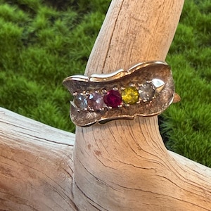 May include: A gold-toned ring featuring a textured, leaf-like metal band set with five round, multi-colored gemstones in a row. The stones include a light blue, a pink, a deep red, a yellow, and a clear white gemstone arranged horizontally.