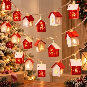 May include: A string of decorative Christmas lights featuring small house-shaped lanterns. Each lantern has a red or white roof and walls with cut-out designs of reindeer and pine trees, illuminated from within to create a warm, festive glow.