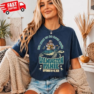 May include: A dark blue t-shirt featuring a graphic of an astronaut playing a guitar while riding a sea turtle. The text reads It's good to be back again, WILMINGTON PANIC, and SEPTEMBER 18+19, 2026, with small flamingo illustrations on the sides.