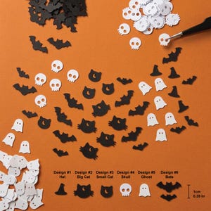 Pode incluir: Uma coleção de pequenas formas de papel cortado, planas, em preto e branco, com desenhos de morcegos, caveiras, fantasmas, chapéus e gatos. Uma ferramenta preta segura uma caveira branca. Uma escala indica um tamanho de 1 cm.