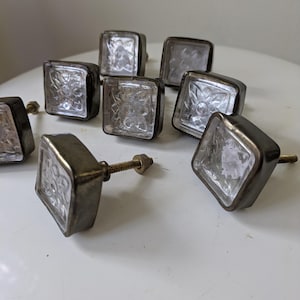 May include: A collection of eight square cabinet knobs featuring a clear, textured glass front with a floral pattern set into a dark, metallic frame. Each knob includes a threaded metal bolt and a small nut attached to the back for installation.