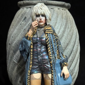 Può includere: Una statuetta in resina dipinta a mano raffigurante una persona con capelli biondi, che indossa una giacca con stampa tigrata sopra una tuta scura iridescente, calze color carne e stivali grigi. La figura tiene una borsa blu su una base.