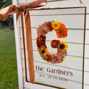 May include: A rectangular glass sign with a gold metal frame, suspended by a gold chain and adorned with a copper-colored velvet ribbon. The sign features a large letter G formed from pressed flowers in shades of orange, yellow, and pink, with the text The Gardners Est. 10.10.2026 printed below.