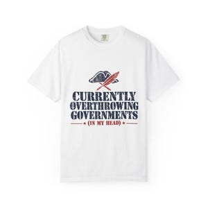 May include: A white short-sleeved t-shirt featuring a distressed graphic of a tricorn hat and a quill pen. Below the graphic, the text reads CURRENTLY OVERTHROWING GOVERNMENTS (IN MY HEAD) in dark blue and red with two small stars on each side.