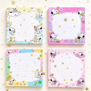 May include: Four square memo pads featuring Snoopy and Woodstock illustrations. The designs include pastel colors, lined writing areas, and various scenes with balloons, boats, and hearts, all decorated with a gold star pattern overlay.
