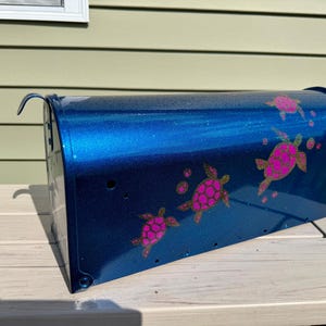 May include: A metallic blue mailbox featuring a glittery finish and a decorative pattern of multiple pink sea turtles. The mailbox has a curved top, a side-mounted metal flag, and a series of small circular holes along the bottom edge of the side panel.