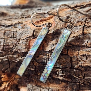 May include: A pair of drop earrings featuring long, rectangular metal bars inlaid with iridescent abalone shell. The shell displays swirling patterns of blue, green, and purple hues. Each earring is attached to a curved metal ear wire hook.