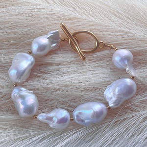 May include: A bracelet featuring seven irregular, lustrous white baroque pearls strung together with small gold-toned beads. The piece is secured with a gold-toned toggle clasp consisting of a circular ring and a T-bar closure mechanism.