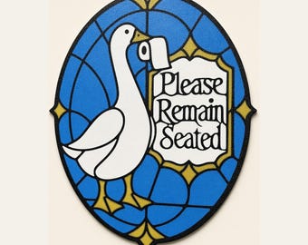 Op de afbeelding: Een ovaal wandbord met een witte gans die een rol toiletpapier in de snavel houdt. De achtergrond is een blauw glas-in-loodpatroon met gouden accenten. Het bord bevat een wit plaatje met de zwarte tekst Please Remain Seated.