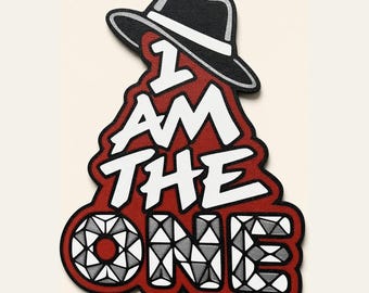 Op de afbeelding: Een stoffen embleem met een zwarte fedora hoed boven de witte tekst I AM THE ONE. Het woord ONE is gevuld met een geometrisch ruitpatroon. Het ontwerp staat op een donkerrode achtergrond met een dikke zwarte randafwerking.