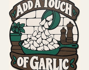 Op de afbeelding: Een decoratief bord met een groene koekenpan gevuld met witte knoflookteentjes die uit een kom worden gegoten. De tekst luidt ADD A TOUCH OF GARLIC. De achtergrond toont een witte bakstenen muur, een groene plant en twee bruine kruidenmolens.