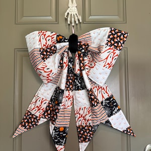 Puede incluir: Un lazo de tela grande con un patrón de retazos de estampados temáticos de Halloween que incluyen fantasmas blancos, calabazas naranjas, telarañas negras, esqueletos y dulces de maíz en una paleta de colores negro, blanco y naranja.