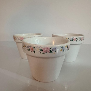 May include: Three white ceramic flower pots with a tapered shape. Each pot features a decorative band around the upper rim, decorated with a colorful floral pattern consisting of small pink roses, blue flowers, and green leaves.