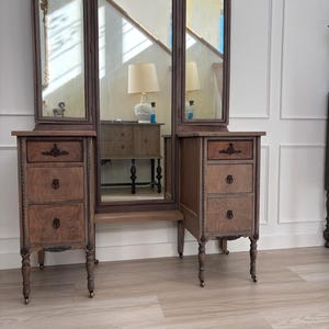 May include: A wooden vanity table featuring a large central mirror flanked by two smaller adjustable side mirrors. The piece includes six drawers with dark metal hardware and is supported by four legs with small metal casters at the base.
