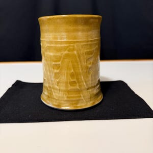 May include: A cylindrical ceramic mug with a mustard yellow glaze and a textured surface featuring vertical, carved, or impressed patterns that create a subtle variation in color depth and light reflection across the exterior of the vessel.