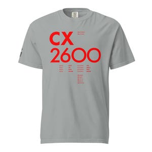 Pode incluir: Uma t-shirt cinzenta clara de manga curta com texto vermelho na parte frontal. O design apresenta o texto grande CX 2600 seguido de especificações técnicas mais pequenas, números de peça e etiquetas como CPU, STELLA e BUFFER na vertical.