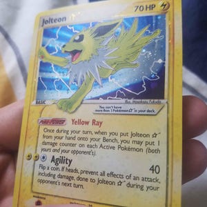 Può includere: Una carta collezionabile Jolteon gialla con 70 HP, caratterizzata da un'illustrazione olografica della creatura elettrica circondata da fulmini. Il testo della carta include il Poke-POWER Raggio Giallo e l'attacco Agilità, con un simbolo a stella.