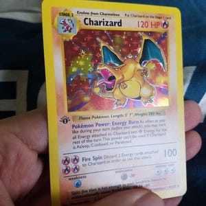May include: A holographic Charizard trading card featuring the Stage 2 Flame Pokémon with 120 HP. The card displays the 1st Edition stamp, the Energy Burn Pokémon Power, and the Fire Spin attack. It lists a length of 5' 7" and weight of 200 lbs.
