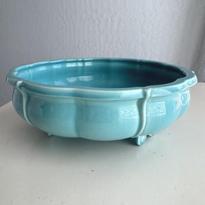 May include: A shallow, round ceramic bowl with a glossy turquoise glaze. The exterior features vertical ridges that create a scalloped edge along the rim. The bowl is supported by small, integrated feet at the base, providing a stable foundation.