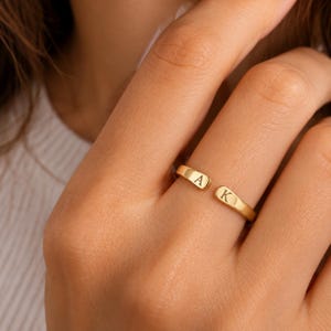 May include: A gold-toned open ring featuring two rounded ends, each engraved with a single capital letter. One end displays the letter A, while the other end displays the letter K. The band has a smooth, polished finish and a simple, adjustable design.