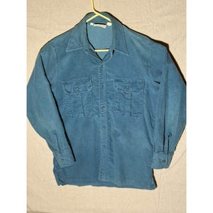 May include: A long-sleeve button-down shirt in a solid teal blue color. The shirt features a pointed collar, a central button placket, and two chest pockets with buttoned flaps. The cuffs also have buttons, and the fabric has a visible corduroy texture.