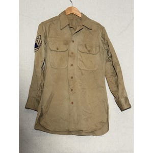 May include: A long-sleeved tan button-down shirt featuring a pointed collar, two chest pockets with buttoned flaps, and a military-style chevron patch on the right sleeve. The shirt is constructed from a solid-colored fabric with a buttoned front.