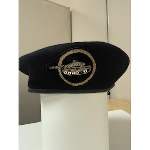 May include: A dark navy blue wool beret featuring a circular embroidered patch on the front. The patch displays a gold-colored tank silhouette enclosed within a circular border, with small metallic accents along the base of the tank design.